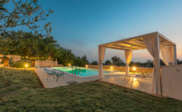 Serene outdoor pool area at dusk featuring a well-lit swimming pool, comfortable seating, and a shaded cabana, surrounded by lush greenery and stone walls, perfect for relaxation and gatherings.