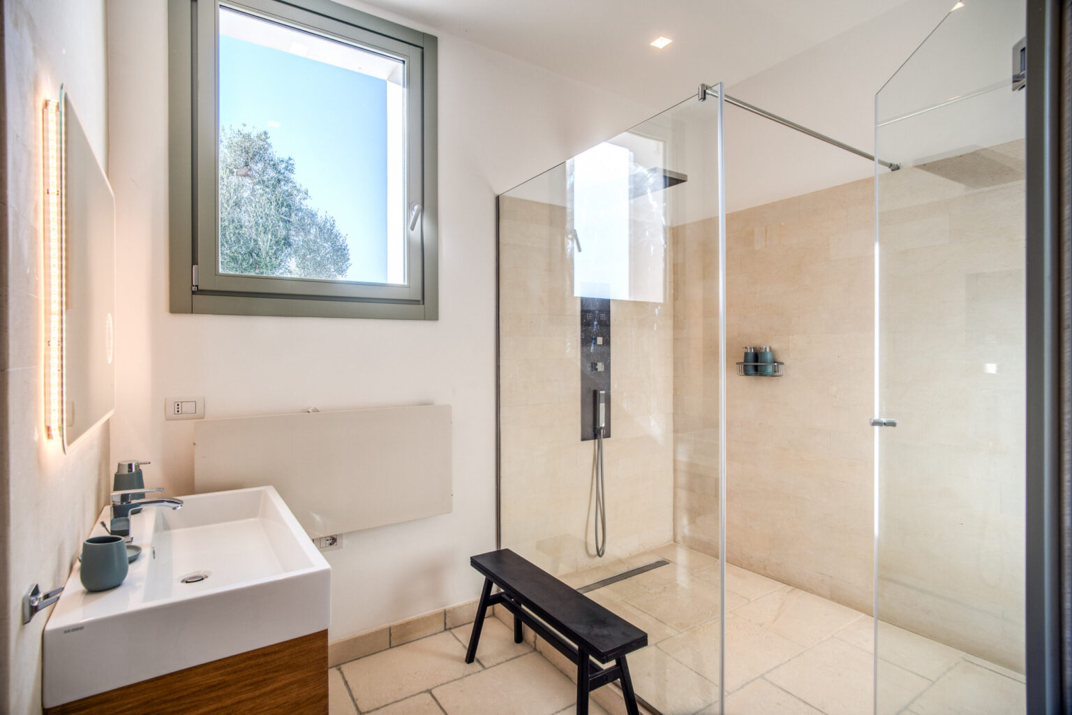 Modern bathroom featuring a glass shower enclosure, minimalist sink with wooden cabinetry, and a stylish black bench. Natural light filters through a large window, illuminating the beige stone tiles and sleek fixtures, creating a serene and contemporary atmosphere. | Only on SopranoVillas