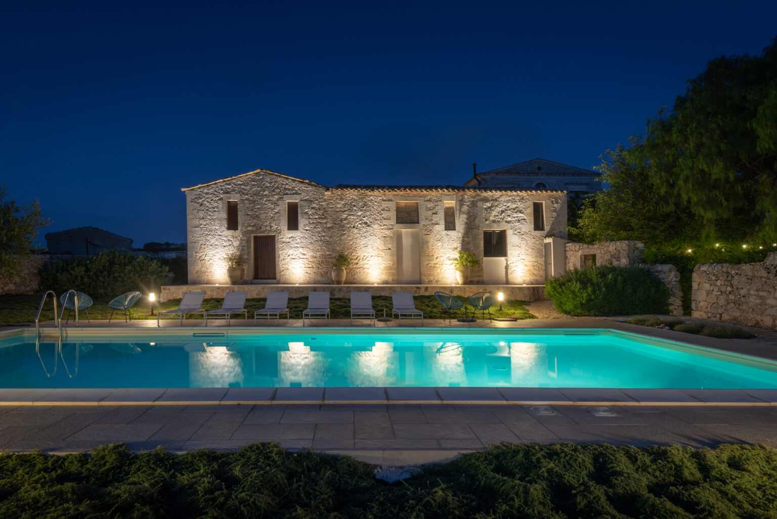 A beautifully lit stone villa at night, featuring a sparkling swimming pool surrounded by lounge chairs and greenery, creating a serene outdoor retreat. | Only on SopranoVillas