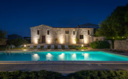 A beautifully lit stone villa at night, featuring a sparkling swimming pool surrounded by lounge chairs and greenery, creating a serene outdoor retreat.