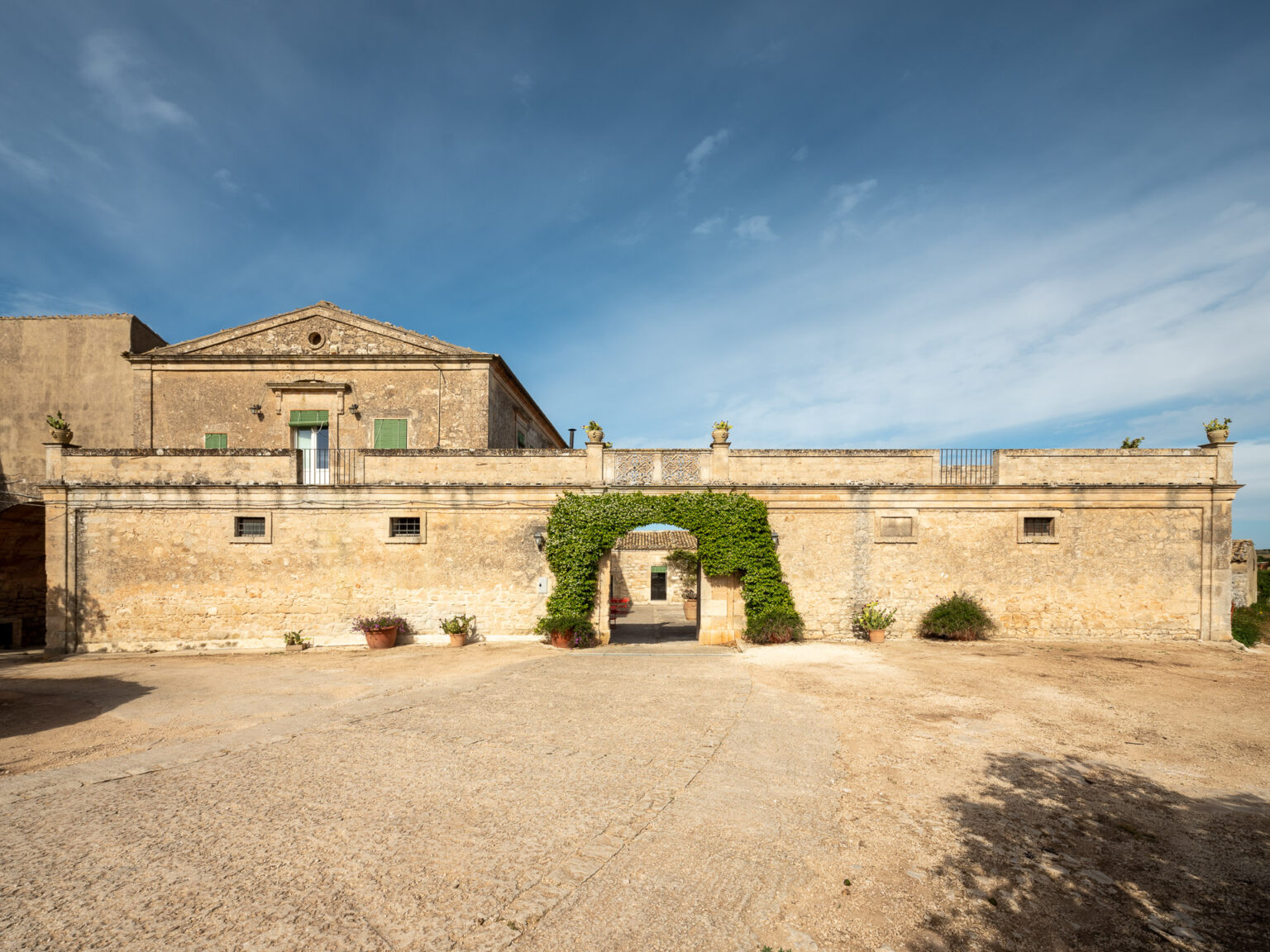 Historic stone building with green shutters and ivy-covered entrance, set against a blue sky, featuring a spacious courtyard and potted plants. Ideal for showcasing architectural heritage and outdoor spaces. | Only on SopranoVillas