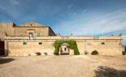 Historic stone building with green shutters and ivy-covered entrance, set against a blue sky, featuring a spacious courtyard and potted plants. Ideal for showcasing architectural heritage and outdoor spaces.