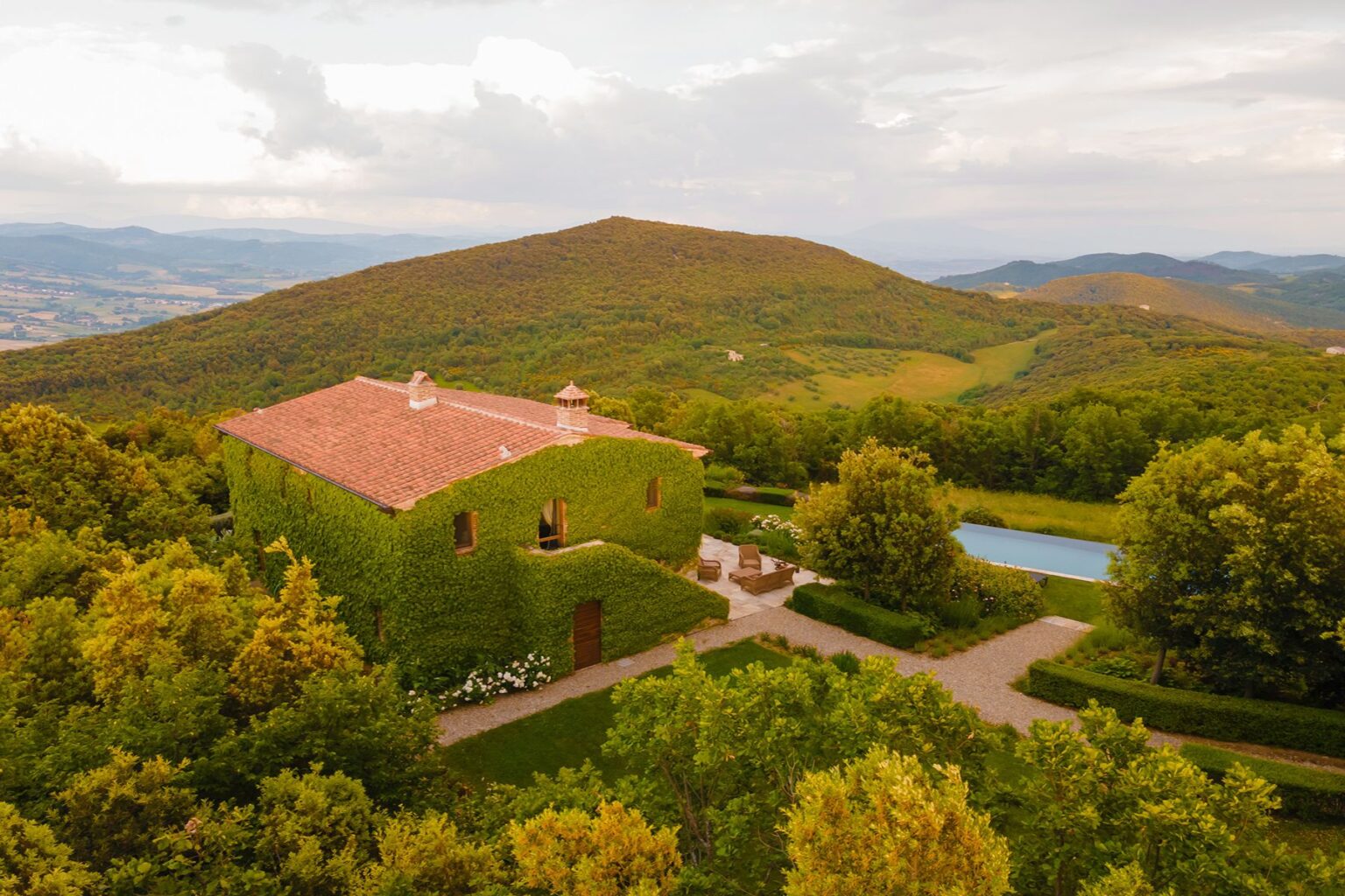 Aerial view of a charming ivy-covered villa surrounded by lush green hills and a serene swimming pool, set against a picturesque landscape in Italy. | Only on SopranoVillas