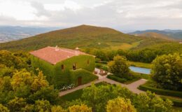 Aerial view of a charming ivy-covered villa surrounded by lush green hills and a serene swimming pool, set against a picturesque landscape in Italy.