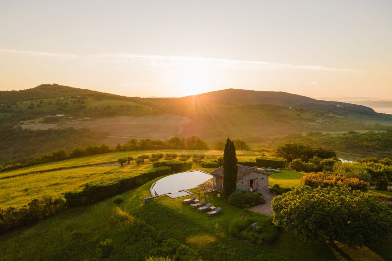 Sunset view over a serene countryside landscape featuring rolling hills, a rustic stone house, and a modern infinity pool surrounded by lush greenery and lounge chairs. Perfect for relaxation and scenic retreats. | Only on SopranoVillas