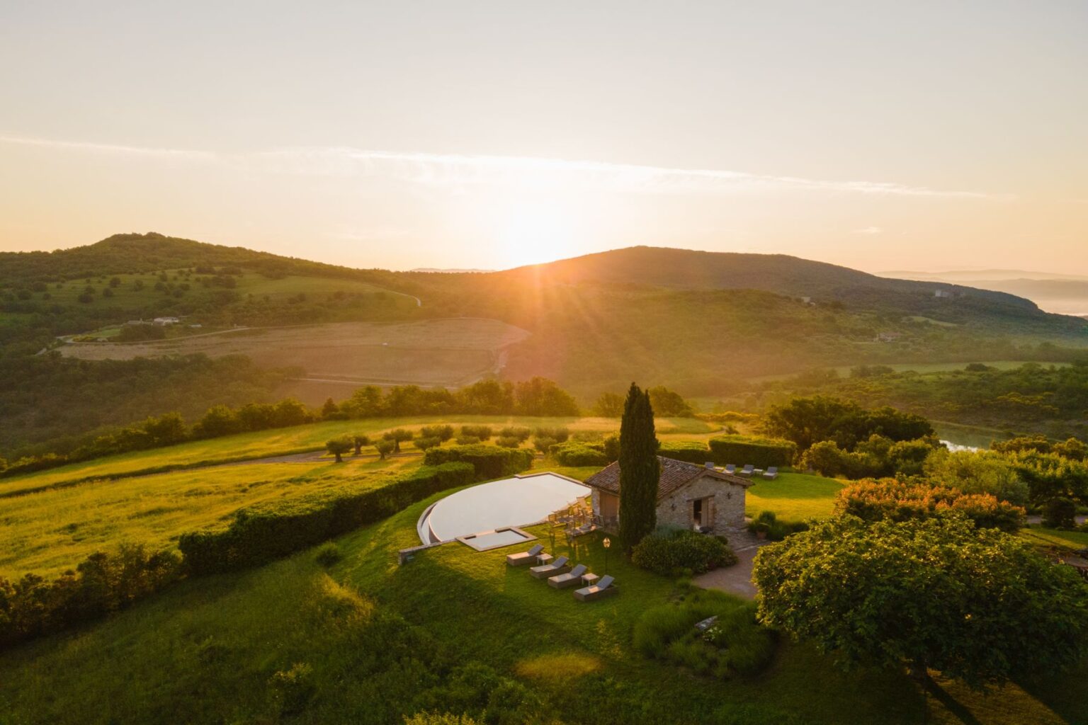 Sunset view over a serene countryside landscape featuring rolling hills, a rustic stone house, and a modern infinity pool surrounded by lush greenery and lounge chairs. Perfect for relaxation and scenic retreats. | Only on SopranoVillas