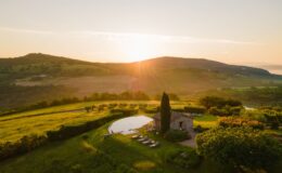 Sunset view over a serene countryside landscape featuring rolling hills, a rustic stone house, and a modern infinity pool surrounded by lush greenery and lounge chairs. Perfect for relaxation and scenic retreats.