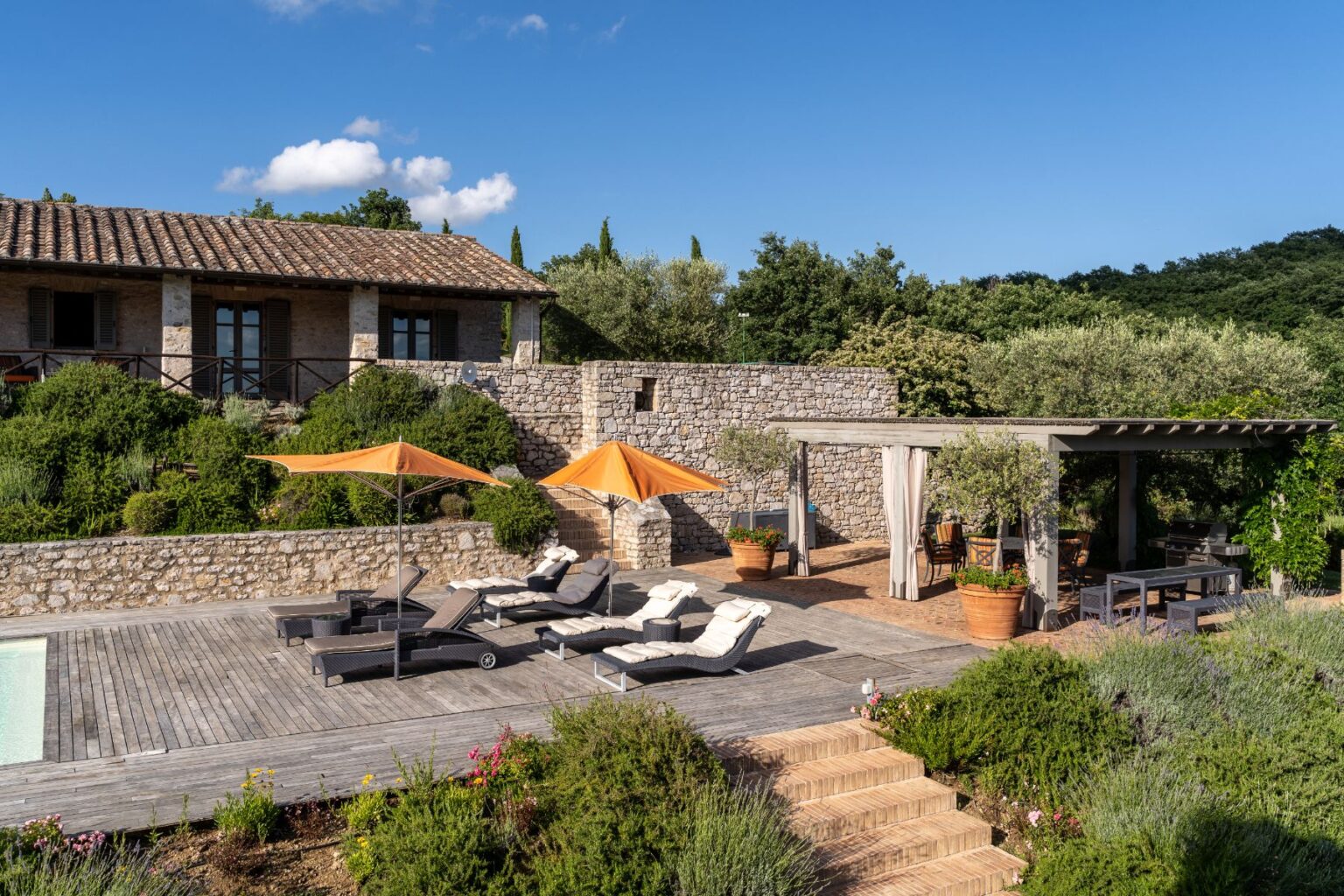 Scenic view of a rustic stone villa with a wooden deck featuring lounge chairs and orange umbrellas, surrounded by lush greenery and flowering plants, under a clear blue sky. The outdoor dining area is partially shaded by a pergola, creating a tranquil and inviting atmosphere. | Only on SopranoVillas