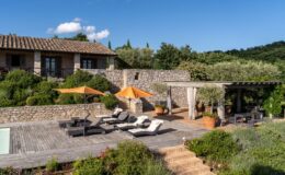 Scenic view of a rustic stone villa with a wooden deck featuring lounge chairs and orange umbrellas, surrounded by lush greenery and flowering plants, under a clear blue sky. The outdoor dining area is partially shaded by a pergola, creating a tranquil and inviting atmosphere.