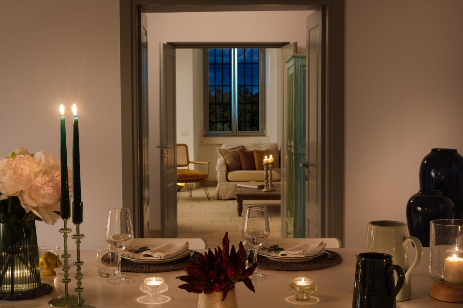 Elegant dining setup with candles and flowers, showcasing a cozy living room in the background with soft lighting and a view of evening skies. Perfect for intimate gatherings and home decor inspiration. | Only on SopranoVillas