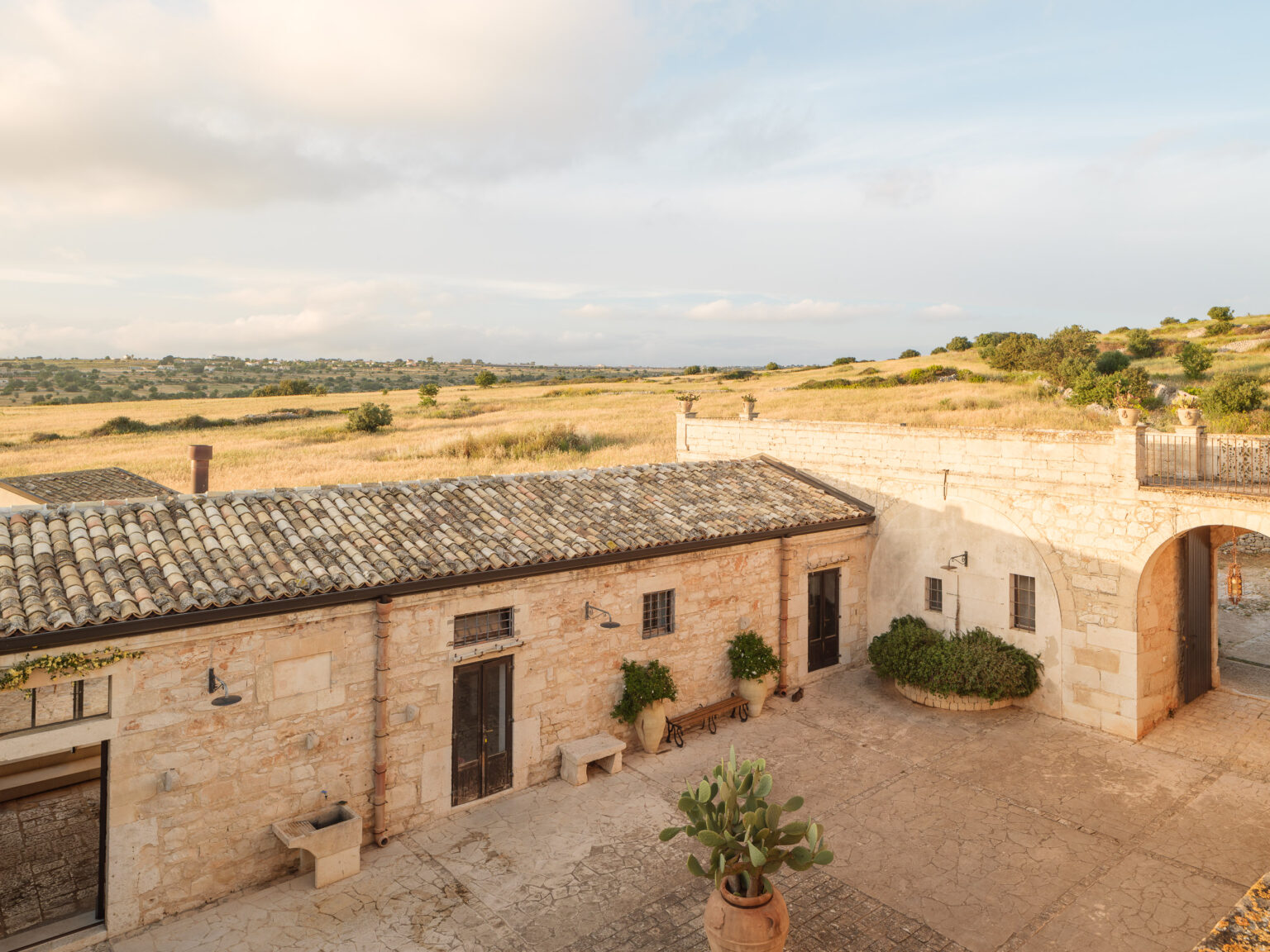 Scenic view of a rustic stone building with a tiled roof, surrounded by lush green fields and rolling hills under a partly cloudy sky. The image captures the serene landscape and architectural charm of a countryside setting. | Only on SopranoVillas