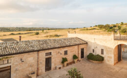 Scenic view of a rustic stone building with a tiled roof, surrounded by lush green fields and rolling hills under a partly cloudy sky. The image captures the serene landscape and architectural charm of a countryside setting.