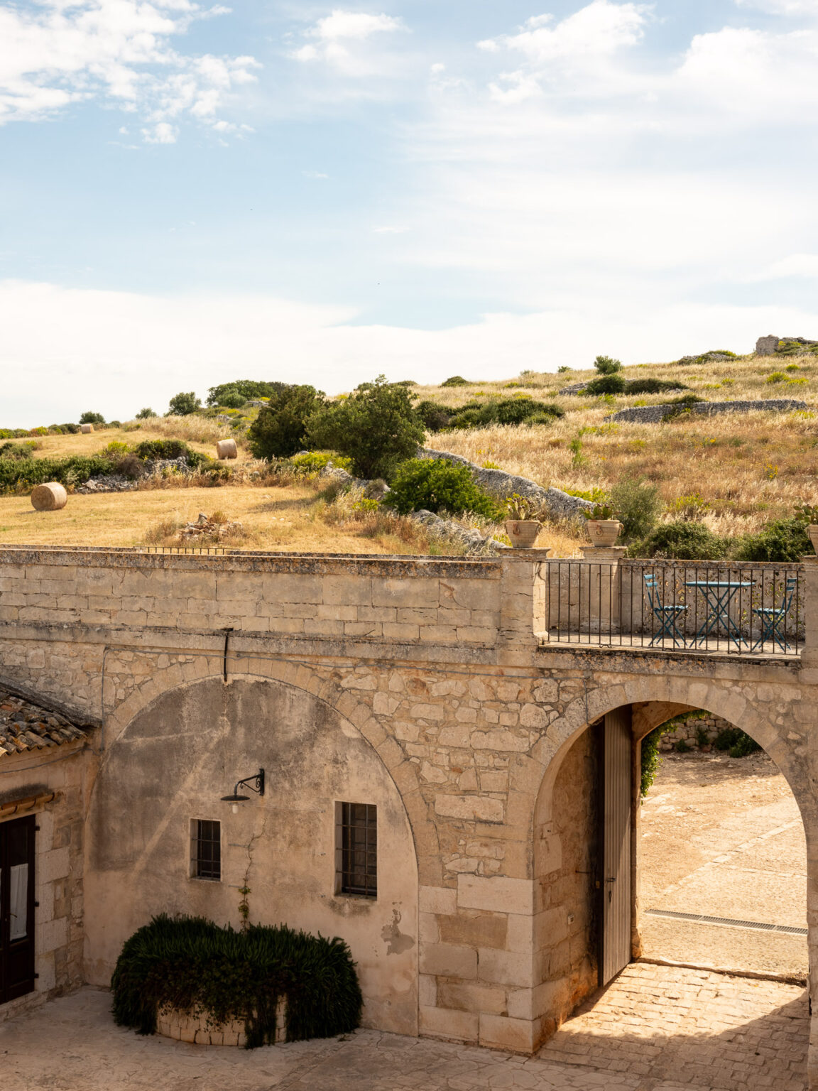 Historic stone archway leading to a scenic landscape of rolling hills and golden fields under a clear blue sky, featuring greenery and rustic stone structures. | Only on SopranoVillas