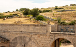 Historic stone archway leading to a scenic landscape of rolling hills and golden fields under a clear blue sky, featuring greenery and rustic stone structures.