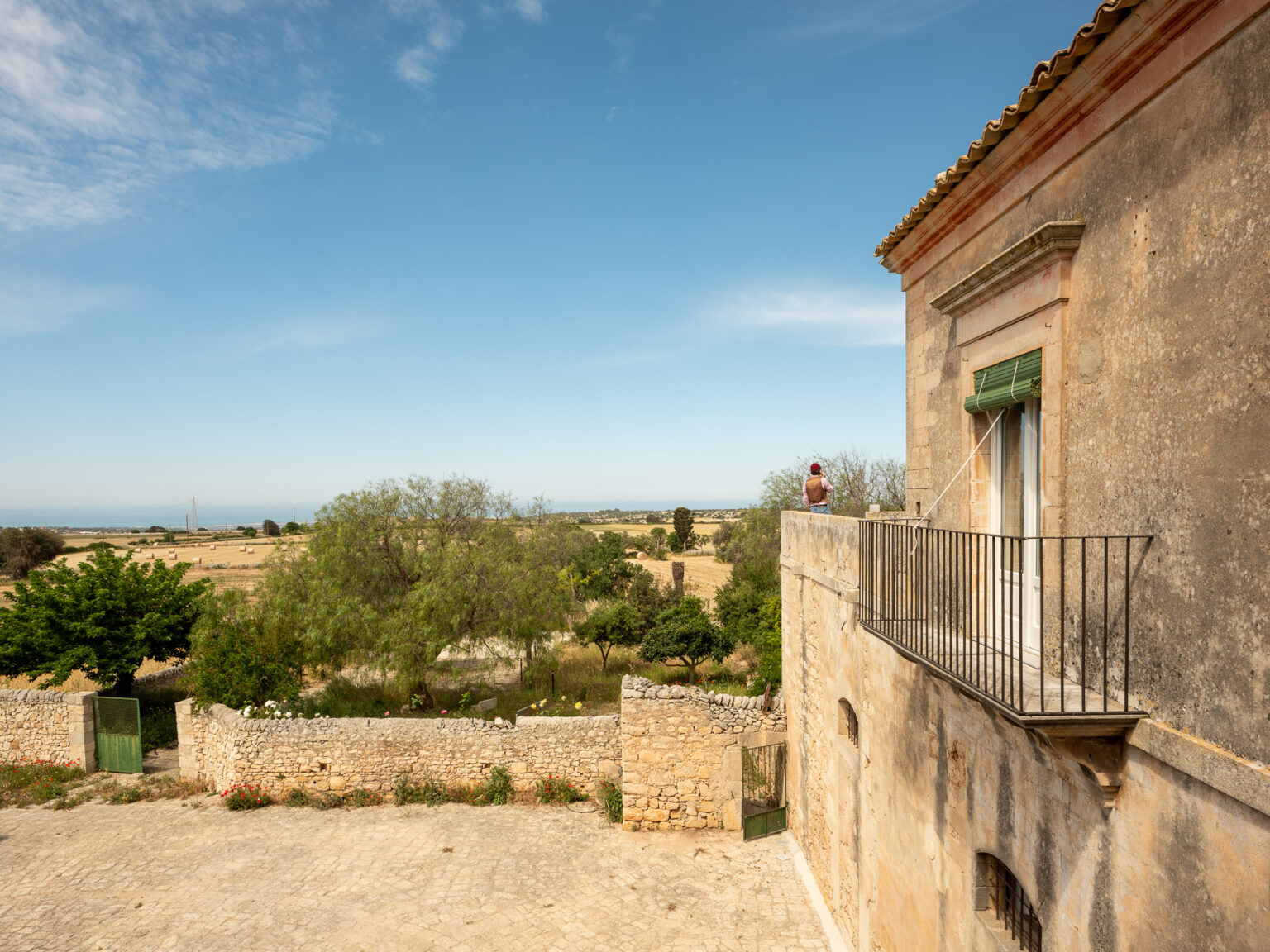 A person enjoying the view from a balcony of an old stone building, overlooking a picturesque landscape of fields and trees under a clear blue sky. | Only on SopranoVillas