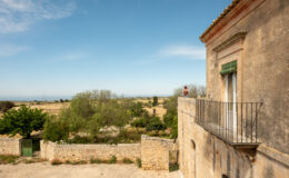A person enjoying the view from a balcony of an old stone building, overlooking a picturesque landscape of fields and trees under a clear blue sky.