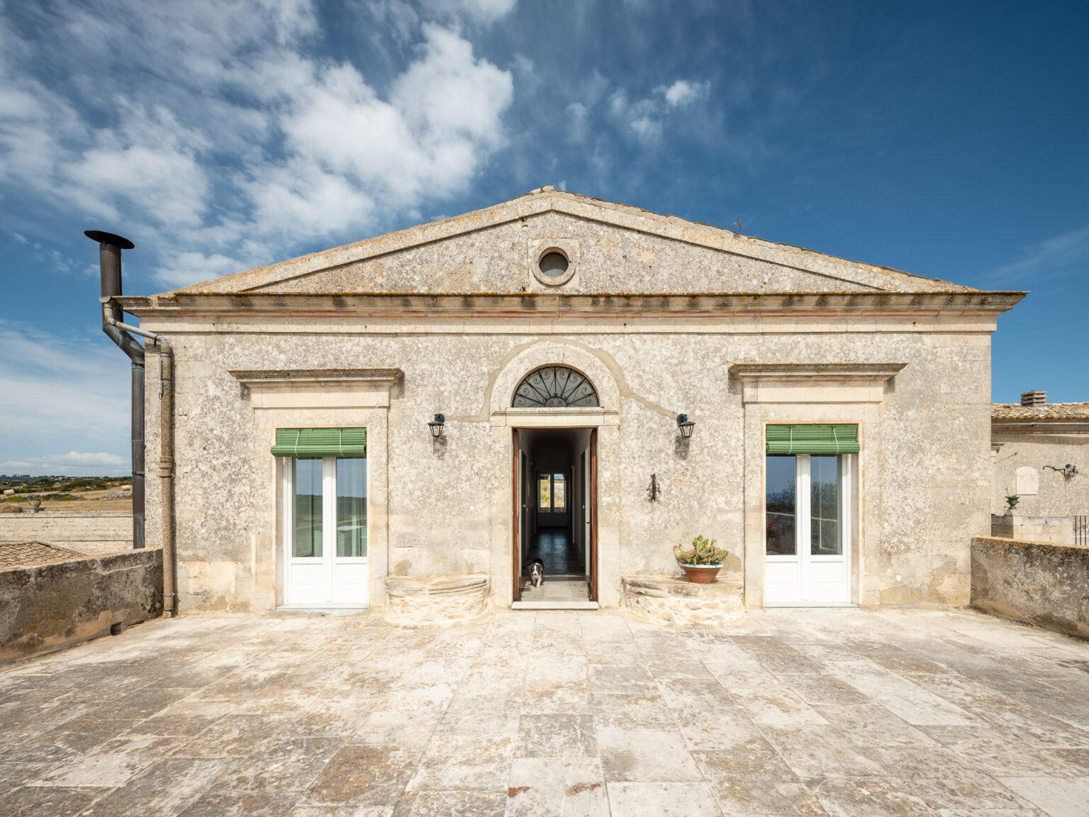 Historic stone building with a classic facade, featuring green shutters and a welcoming entrance. A sunny terrace is visible, surrounded by scenic landscapes under a blue sky with clouds. | Only on SopranoVillas