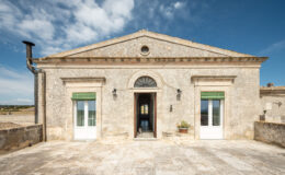 Historic stone building with a classic facade, featuring green shutters and a welcoming entrance. A sunny terrace is visible, surrounded by scenic landscapes under a blue sky with clouds.