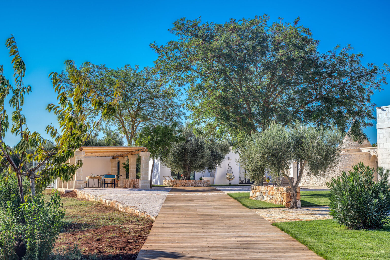 Scenic outdoor pathway leading to a modern stone structure surrounded by lush greenery and trees, featuring a shaded seating area with a table, ideal for relaxation and outdoor dining. | Only on SopranoVillas