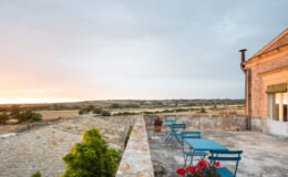 Scenic view from a terrace featuring blue metal chairs and tables, overlooking lush fields and a sunset sky in a rural landscape.