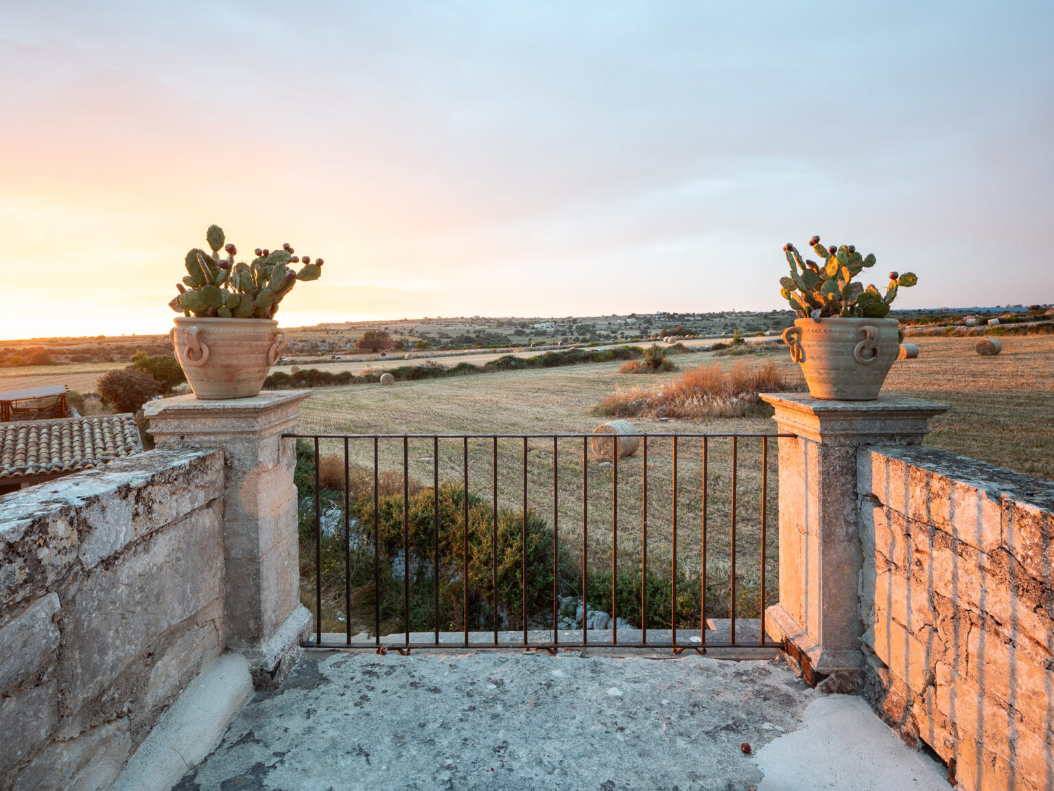 Scenic view of a rustic stone balcony with potted cacti, overlooking a vast golden field at sunset, framed by a black metal gate. The serene landscape features hay bales in the distance under a colorful sky. | Only on SopranoVillas