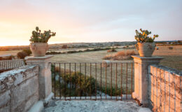 Scenic view of a rustic stone balcony with potted cacti, overlooking a vast golden field at sunset, framed by a black metal gate. The serene landscape features hay bales in the distance under a colorful sky.