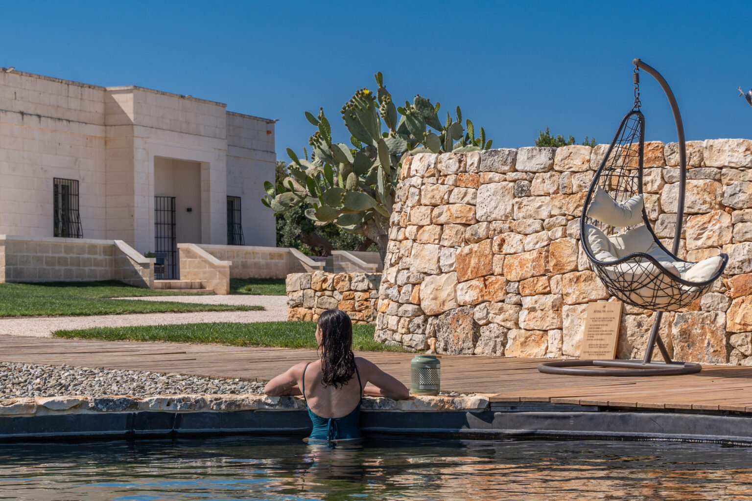 Person relaxing in a swimming pool at a modern outdoor space with stone walls, a hanging chair, and cacti under a clear blue sky. | Only on SopranoVillas