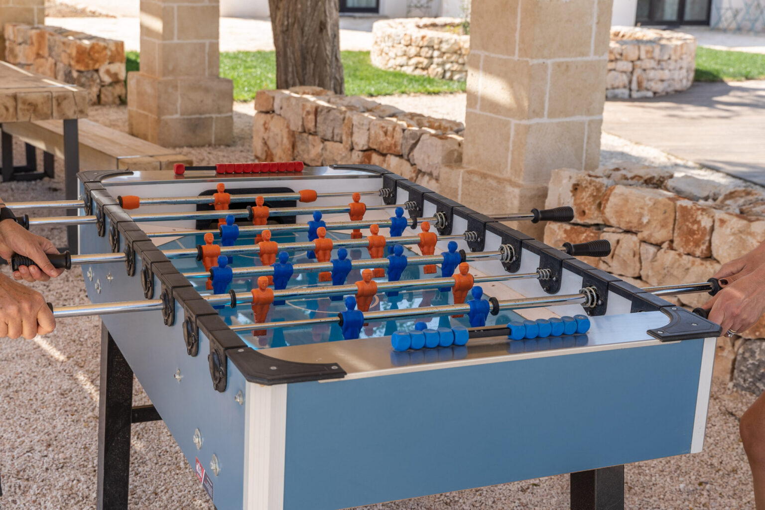Two players engaging in a competitive game of foosball at an outdoor table, showcasing colorful player figurines in blue and orange on a well-maintained foosball table. | Only on SopranoVillas
