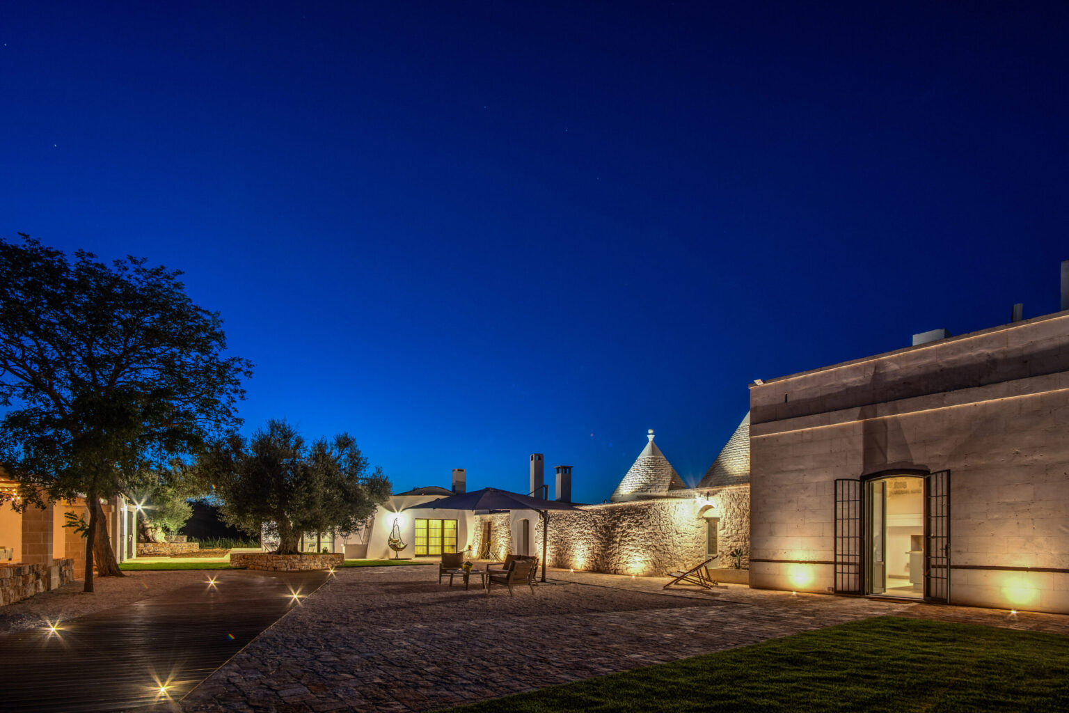 Tranquil evening view of a traditional trullo house and surrounding buildings, illuminated by soft outdoor lighting against a deep blue sky. Ideal for showcasing unique architectural styles and serene outdoor spaces. | Only on SopranoVillas