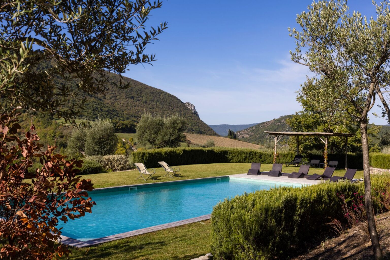 Scenic view of a tranquil swimming pool surrounded by lush greenery and mountains, featuring lounge chairs and a shaded pergola, ideal for relaxation and outdoor leisure. | Only on SopranoVillas
