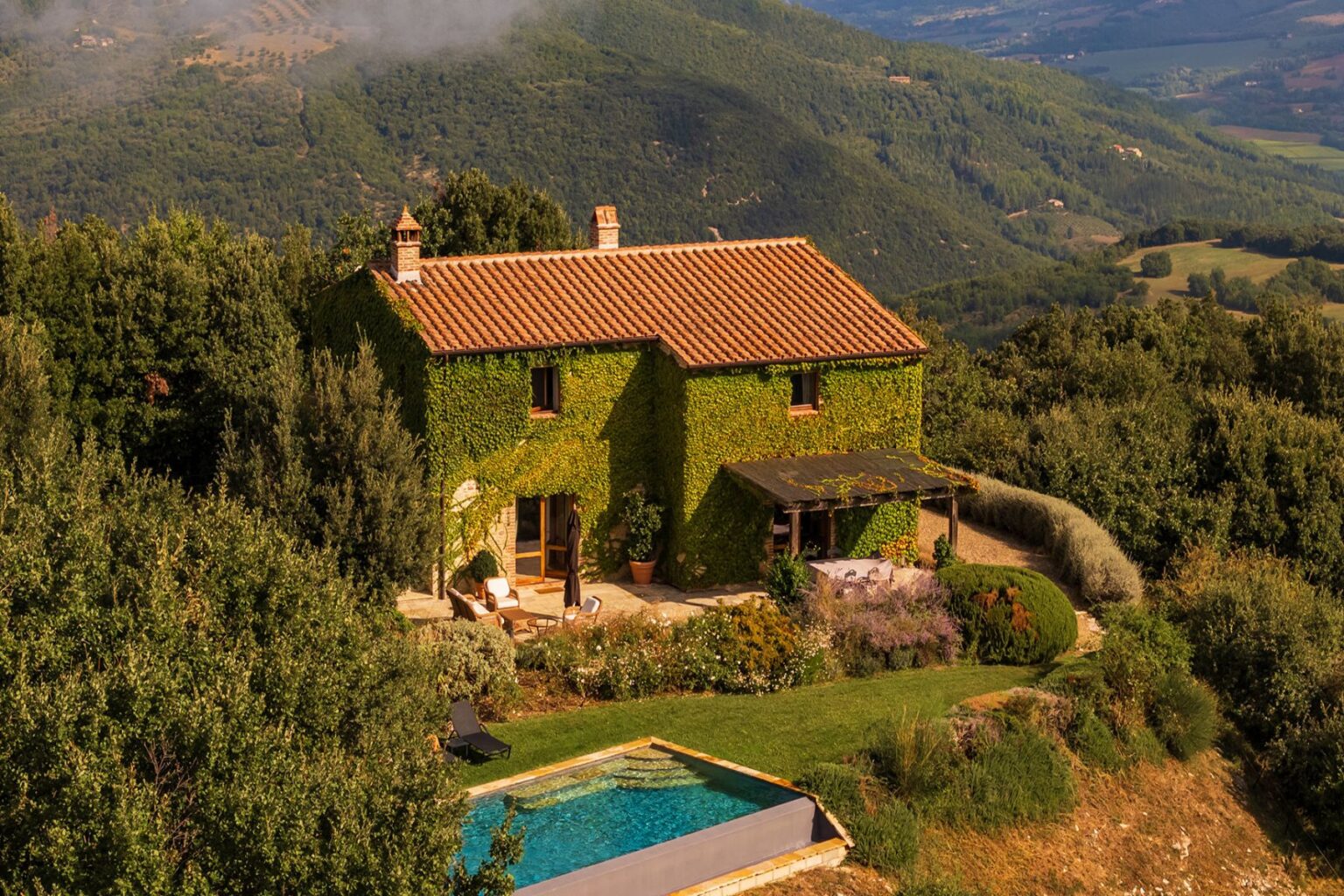 Charming countryside villa covered in lush green ivy, featuring a swimming pool and surrounded by vibrant gardens, set against a backdrop of rolling hills and mountains. | Only on SopranoVillas