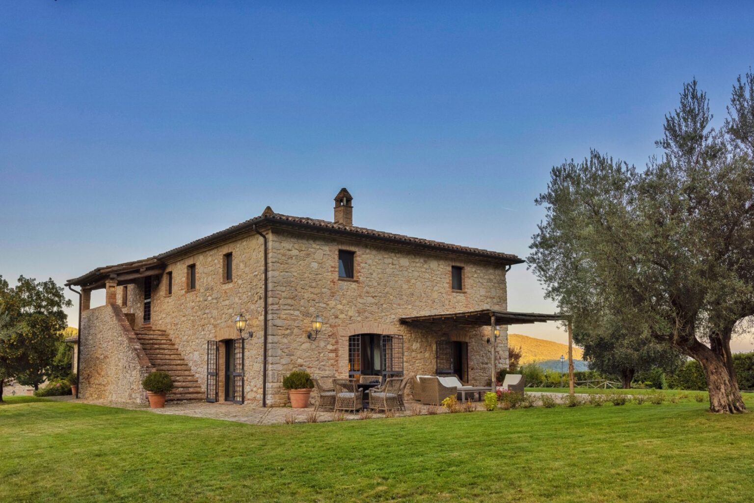 Charming stone farmhouse with a lush green lawn, outdoor seating area, and scenic landscape in the background, ideal for countryside retreats. | Only on SopranoVillas