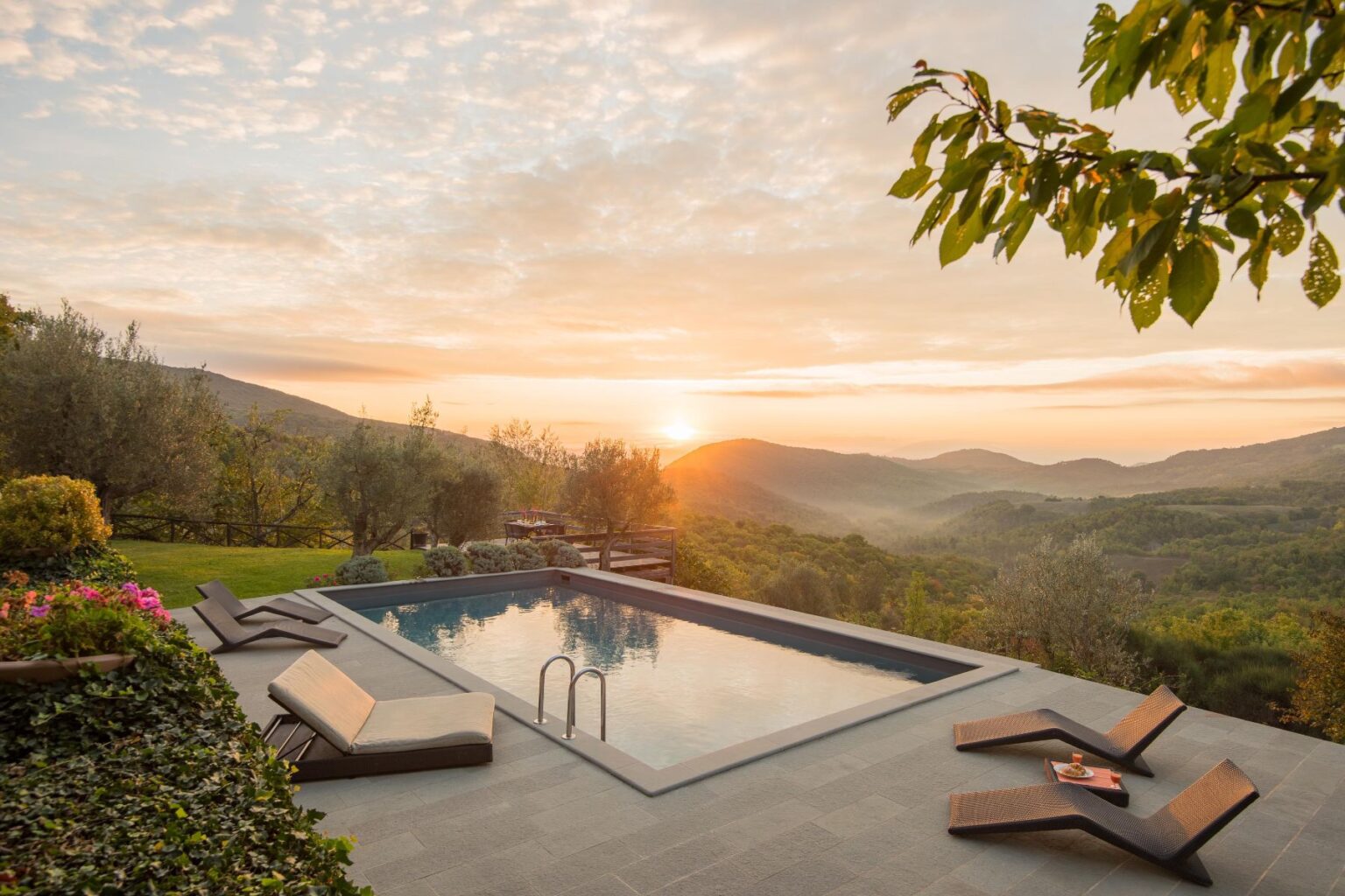 Luxurious outdoor pool area with modern lounge chairs, surrounded by lush greenery and olive trees, showcasing a breathtaking sunset over rolling hills. Ideal for relaxation and enjoying nature. | Only on SopranoVillas