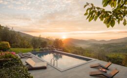 Luxurious outdoor pool area with modern lounge chairs, surrounded by lush greenery and olive trees, showcasing a breathtaking sunset over rolling hills. Ideal for relaxation and enjoying nature.