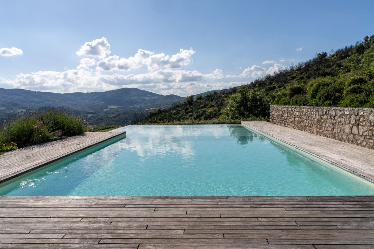 Serene infinity pool overlooking rolling hills and mountains under a partly cloudy sky, featuring a wooden deck and lush greenery. Ideal setting for relaxation and outdoor living. | Only on SopranoVillas