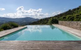 Serene infinity pool overlooking rolling hills and mountains under a partly cloudy sky, featuring a wooden deck and lush greenery. Ideal setting for relaxation and outdoor living.