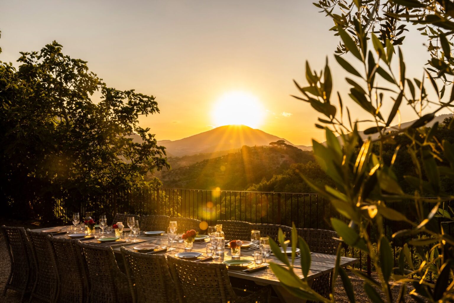 Sunset dining setup with a beautifully arranged table featuring elegant glassware and floral centerpieces, overlooking a picturesque mountain landscape bathed in golden light. Lush greenery frames the scene, creating an inviting atmosphere for outdoor gatherings. | Only on SopranoVillas