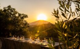 Sunset dining setup with a beautifully arranged table featuring elegant glassware and floral centerpieces, overlooking a picturesque mountain landscape bathed in golden light. Lush greenery frames the scene, creating an inviting atmosphere for outdoor gatherings.