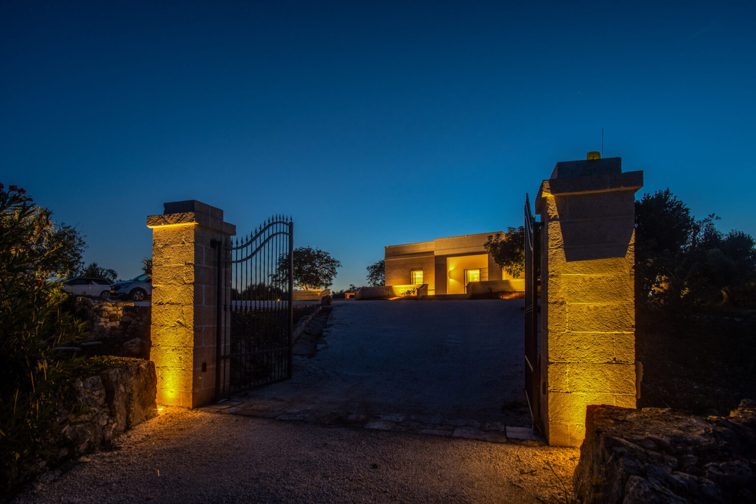 Elegant entrance gate illuminated at dusk, leading to a modern home surrounded by lush greenery and a serene landscape. Ideal for showcasing luxury real estate or tranquil living spaces. | Only on SopranoVillas