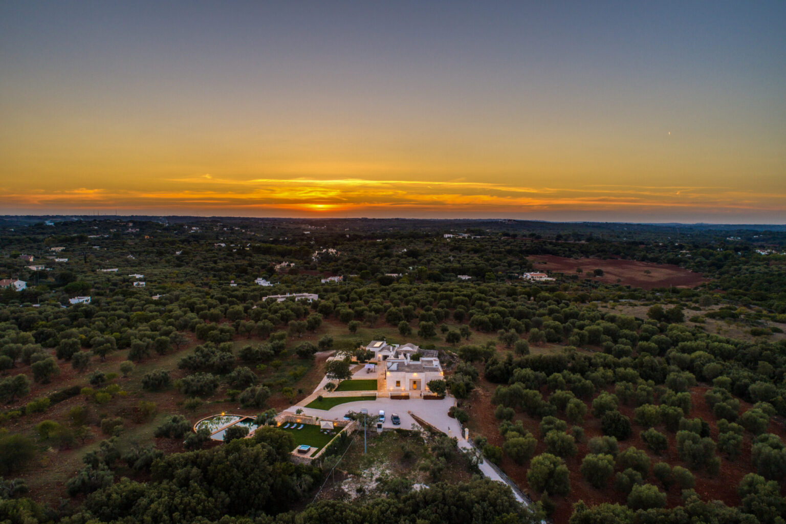 Aerial view of a serene landscape featuring a modern villa surrounded by olive trees at sunset, showcasing vibrant orange and purple hues in the sky. | Only on SopranoVillas