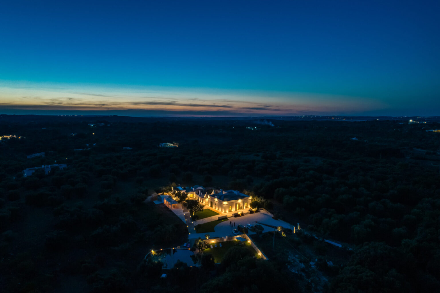 Aerial view of a beautifully lit property surrounded by lush greenery at dusk, showcasing a stunning sunset over the horizon. | Only on SopranoVillas