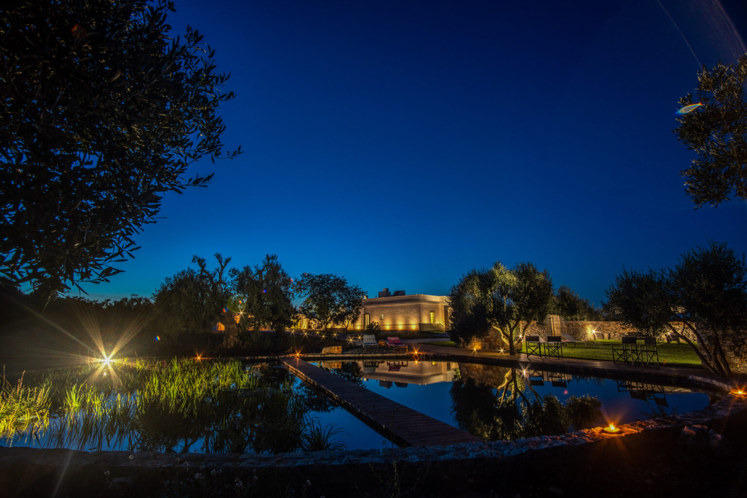 Serene night view of a landscaped garden featuring a tranquil pond, illuminated by soft lights, with a modern building in the background against a deep blue sky. | Only on SopranoVillas