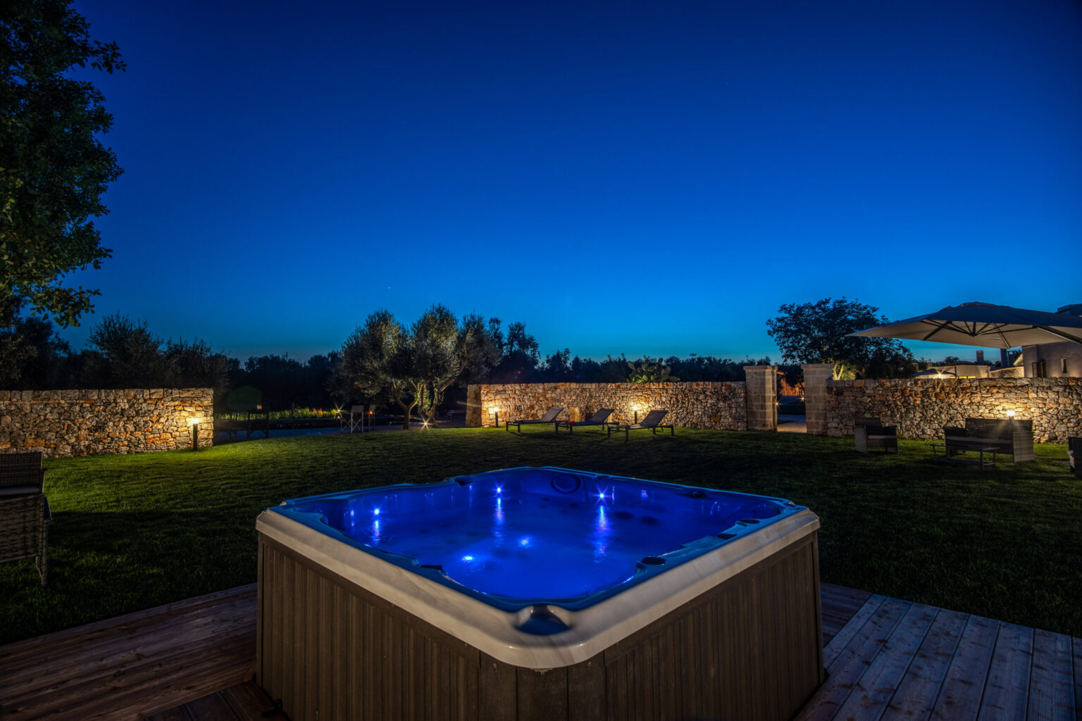 Hot tub illuminated at night in a serene outdoor setting with lush greenery and stone walls, perfect for relaxation and evening gatherings. | Only on SopranoVillas
