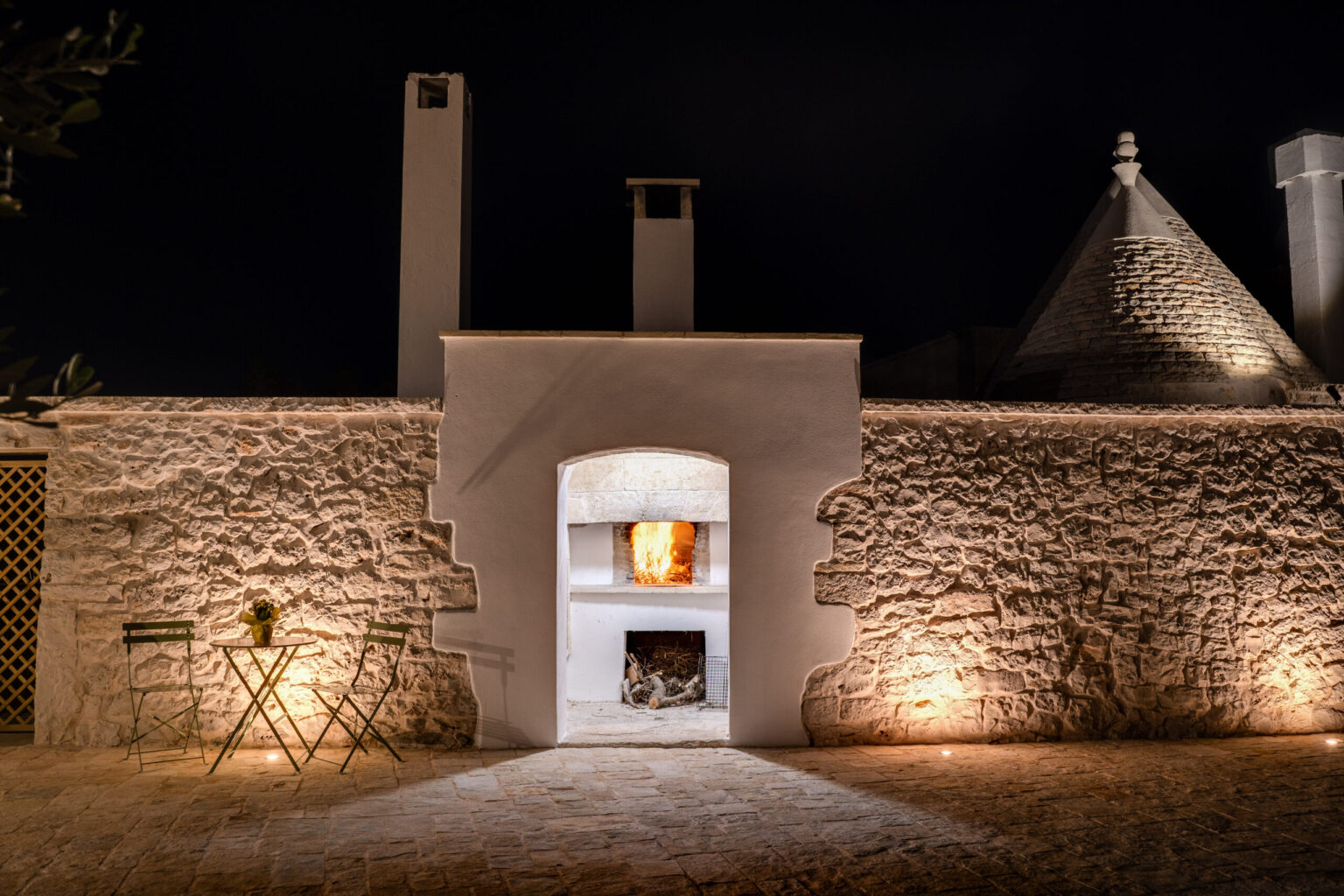 Traditional stone trullo house at night with illuminated outdoor fireplace and cozy seating area, showcasing rustic architecture and tranquil ambiance. | Only on SopranoVillas