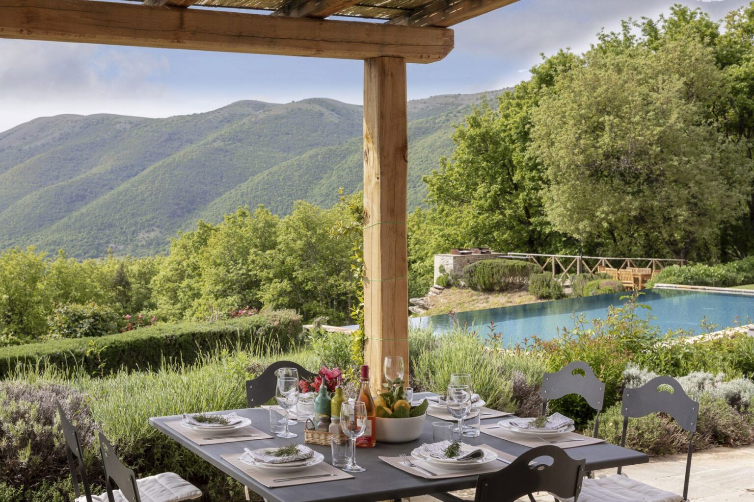 Scenic outdoor dining area with a table set for a meal, featuring elegant tableware and a centerpiece of fresh fruits. In the background, lush green hills and a serene swimming pool create a tranquil atmosphere, ideal for relaxation and gatherings. | Only on SopranoVillas