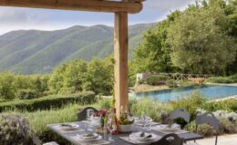 Scenic outdoor dining area with a table set for a meal, featuring elegant tableware and a centerpiece of fresh fruits. In the background, lush green hills and a serene swimming pool create a tranquil atmosphere, ideal for relaxation and gatherings.