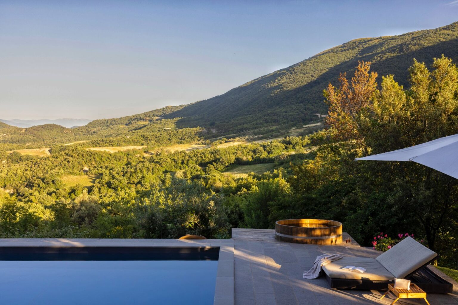 Luxurious outdoor pool area with a stunning mountain view, featuring a wooden hot tub and comfortable lounge chair, surrounded by lush greenery under a clear sky. Perfect for relaxation and enjoying nature. | Only on SopranoVillas