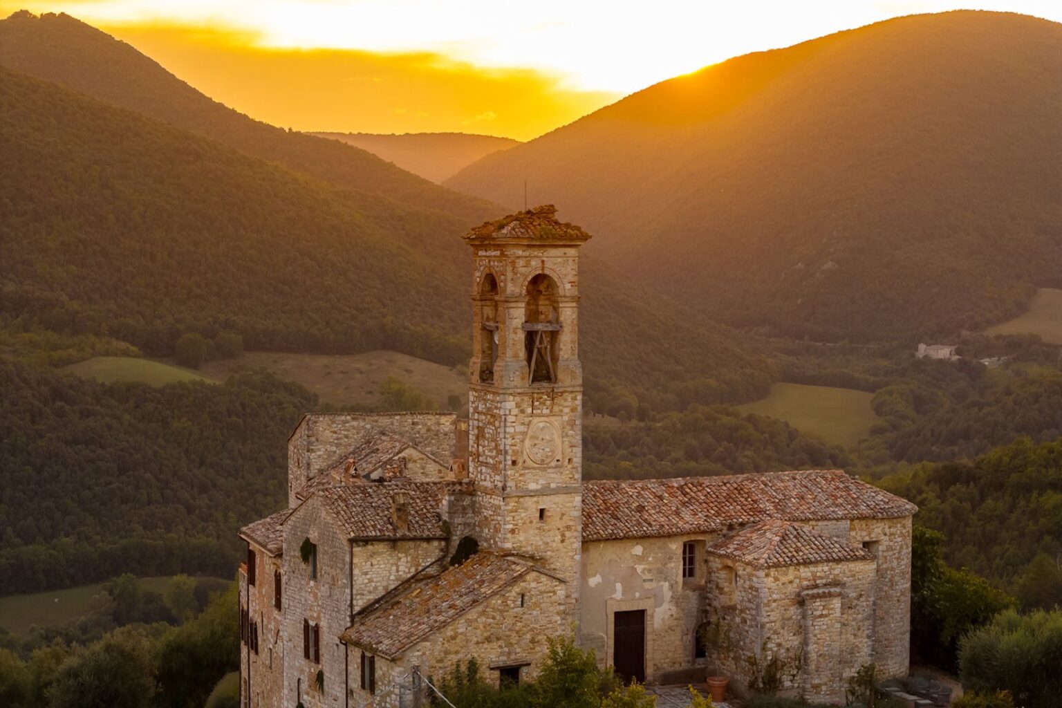 Historic stone building with a bell tower at sunset, surrounded by rolling green hills and mountains in the background. | Only on SopranoVillas