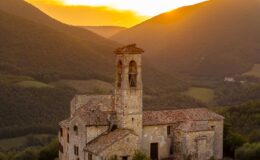 Historic stone building with a bell tower at sunset, surrounded by rolling green hills and mountains in the background.