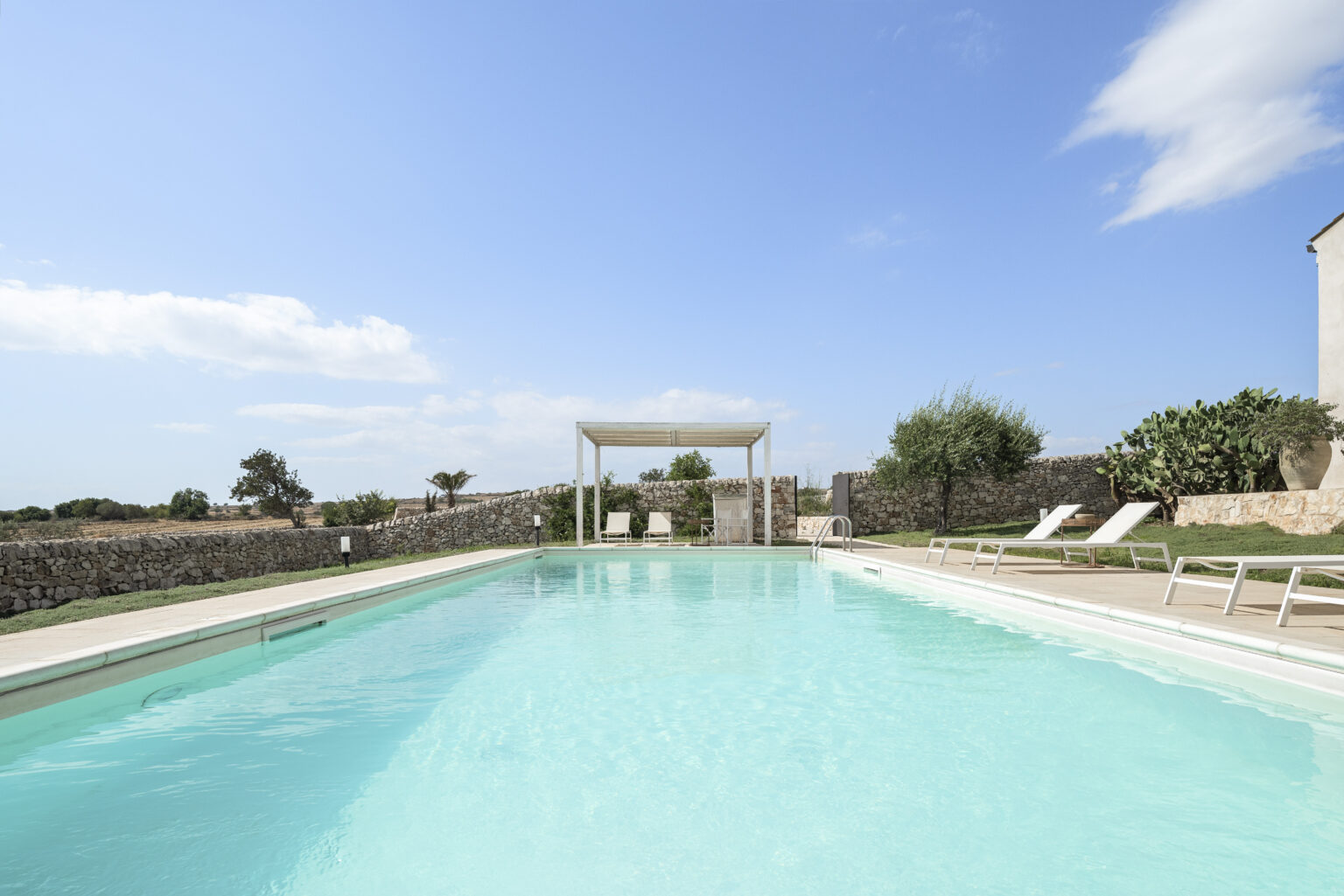 Serene outdoor swimming pool surrounded by lush greenery and a stone wall, featuring lounge chairs and a shaded pergola under a clear blue sky. Ideal for relaxation and leisure. | Only on SopranoVillas